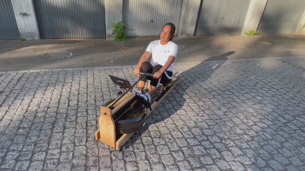 Video: Frau rudert im Freien auf dem TWHEELS Wasserrudergerät aus Eichenholz, mit Rudersitz und Wassertank im Bild.